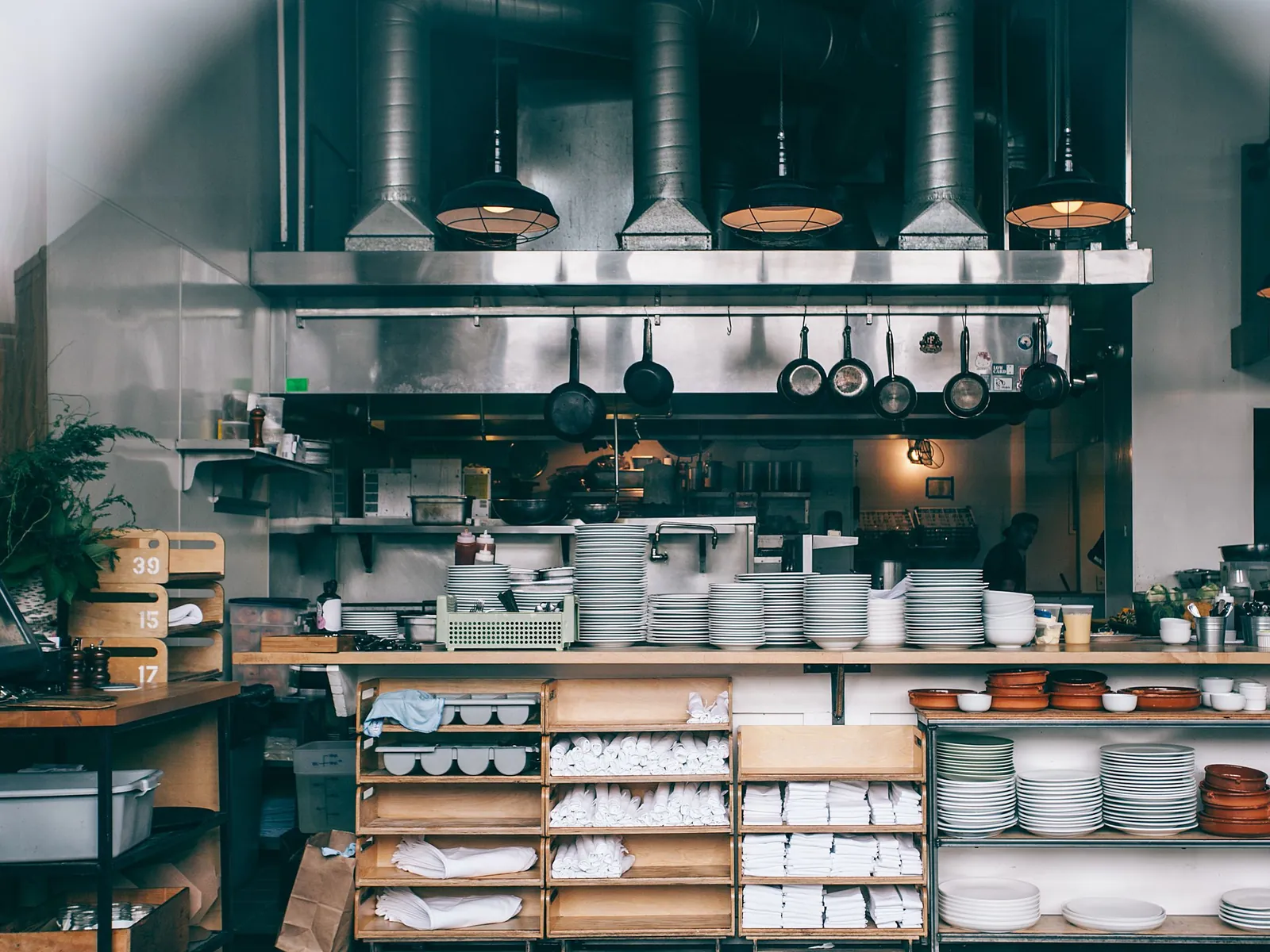 The pass of a working commercial kitchen: stacked plates and bowls, folded linen on open shelving, and pans hanging beneath the extraction hood.