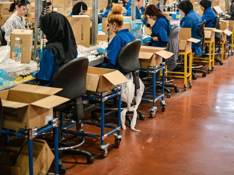 Workers in blue uniforms seated along a factory assembly line, packing finished goods into cardboard cartons.