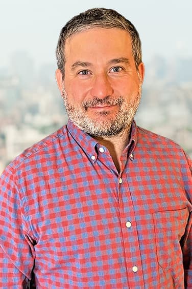 Brian Berger, founding partner, in a red and blue checked shirt, smiling, with a hazy city skyline behind him.