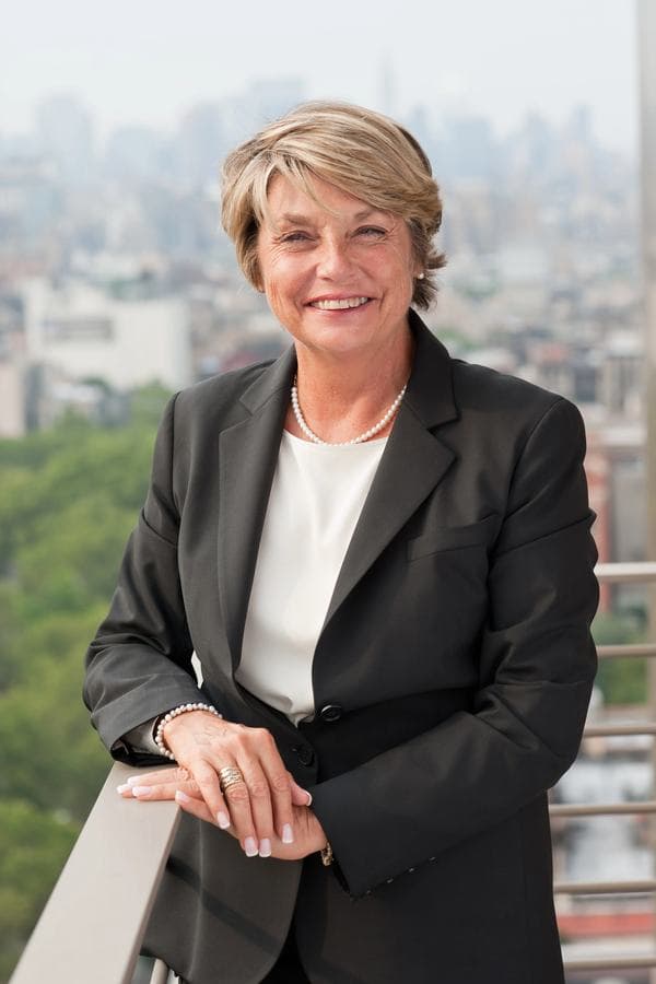 Lynne Jacoby, founding partner, in a dark jacket, leaning on a terrace rail with a city skyline behind her.